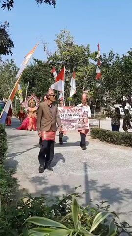 A group of people are participating in a parade. In the foreground, two men walk side-by-side, each holding a banner. The man on the left wears a brown jacket and a red and black patterned sarong. The man on the right wears a white shirt and a red and black patterned sarong, and a red cap. Behind them, two men in white shirts and red and black patterned sarongs carry Indonesian flags. In the background, a person in an elaborate, colorful costume, possibly a dancer or performer, walks with a train of red fabric. Other individuals, dressed in red and green outfits, are visible further behind. The parade appears to be taking place outdoors on a sunny day, with trees and foliage surrounding a paved pathway. Red and white Indonesian flags are also strung along the path.
