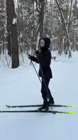 A young woman stands in a snowy forest, facing slightly to the left, wearing a black hooded jacket and pants. She is on cross-country skis with black and bright yellow details. She holds ski poles in her gloved hands, gesturing with them as if she is teaching or explaining something. The ground is covered in snow, and trees are visible in the background. A faint path or clearing is seen under the snow.  The lighting suggests a winter day.