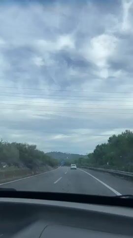 The video shows a first-person perspective from a vehicle traveling on a highway. The road has multiple lanes and is surrounded by vegetation and hills in the distance. The sky is overcast with scattered clouds. Several other vehicles are visible on the road ahead, including a white car in the right lane and a blue sign further down. Power lines are visible strung across the top of the frame. The interior of the vehicle, specifically the dashboard, is partially visible in the foreground.