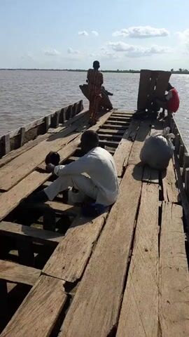 The video shows the interior of a wooden boat on a body of water. Three people are visible. In the foreground, a person wearing a light-colored garment sits facing away from the camera, looking towards the front of the boat. The person's legs are bent, and they are seated on the wooden deck, which is made of planks with gaps between them. To the right of this person, a large, light-colored bag or bundle rests on the deck. Further ahead, two other people are standing. One person, wearing a reddish top and dark bottoms, is near the back of the boat, interacting with something obscured by a wooden structure. Another person, wearing a striped outfit, stands near the front, also appearing to be engaged with something. The boat's wooden structure is weathered and shows signs of age. The water is a murky brown, and the sky is bright with some clouds. The scene suggests a calm day on the water.
