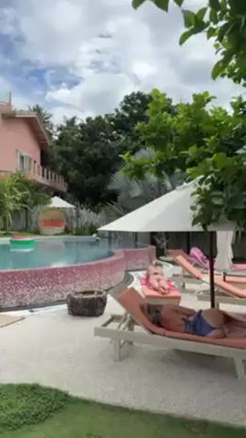 The video shows a swimming pool surrounded by lounge chairs and a pink building in the background. Two women are lying on the lounge chairs, sunbathing. The pool is decorated with pink mosaic tiles, and a watermelon-shaped inflatable is floating in the water. The area around the pool is covered with gravel and grass. There are several white parasols providing shade. The building is a two-story structure with white balconies. The sky is partly cloudy.