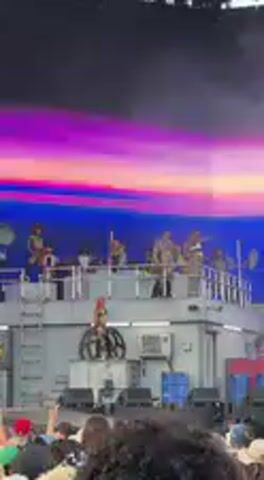 This video depicts a live performance at an outdoor music festival, likely in Indio, United States. A group of performers or DJs are positioned on a distinctive multi-level stage structure that resembles stacked containers or a modular building. Several individuals on stage raise their arms, indicating interaction with the crowd or a moment of excitement during the performance.

Behind the stage, a large screen displays dynamic and vibrant lighting effects, predominantly featuring shades of blue, purple, and pink, which shift and change throughout the clip. At one point, the screen briefly darkens before returning to its colorful display. In the immediate foreground, the heads and shoulders of a large audience are visible, suggesting a crowded event. The lighting conditions, with the emphasis on stage lights and screen visuals, indicate that the event is taking place in the evening or at night. No discernible text is visible on the stage or screen.