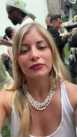 The video shows a blonde woman with blue eyes wearing a white tank top and a triple-strand pearl necklace. She looks at the camera and smiles slightly. She is surrounded by other people, who are seated at tables in an outdoor setting, possibly a restaurant or event. There is greenery and decorations around. The video is shot from a close-up angle, with the woman as the main focus. There is background music playing.