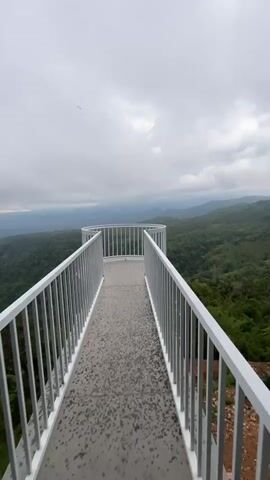 This video showcases a modern, white-railed observation deck offering a panoramic view of a lush, green, mountainous landscape. The deck itself is paved with a textured gray material, appearing somewhat wet, suggesting recent rain or mist. The railing is sleek and minimalist, forming a curved barrier around the edge of the viewpoint. Beyond the railing, rolling hills covered in dense vegetation stretch out to the horizon, fading into a hazy, distant landscape. The sky is overcast with heavy, gray clouds, casting a soft, diffused light over the scene. The overall atmosphere is serene and slightly melancholic, evoking a sense of peaceful solitude amidst nature. The time of day appears to be late afternoon or early morning, given the subdued lighting. While no people are visible, the presence of the constructed viewpoint implies it is a place for visitors to appreciate the scenery. The location context provided is Nam Hải, Vietnam, which aligns with the tropical or subtropical appearance of the dense greenery.