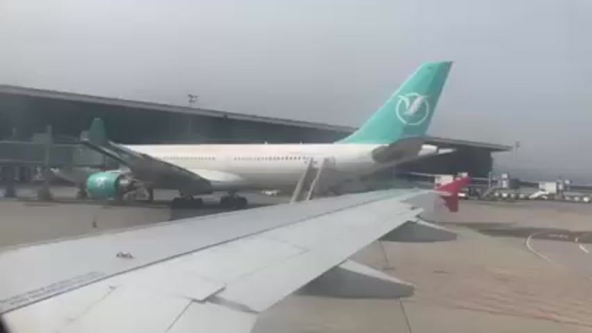 Here is a concise description of the video:

The video shows an airplane at an airport, as viewed from the window of another plane. The plane in the foreground is white with a light grey wing. The plane further away is white with turquoise accents on its tail and engine, and has a staircase leading up to its entrance. An airport terminal building is visible in the background.  The overall scene appears hazy.