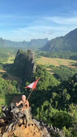 A woman sits on a rocky precipice, holding a flag with red and white stripes. She is wearing a black sports bra and green pants. Behind her, a lush green valley stretches out, dotted with rice paddies and surrounded by jagged, mist-shrouded mountains under a clear blue sky. A road winds through the valley below. The camera angle is from a slightly higher vantage point, looking down at the woman and the expansive landscape.