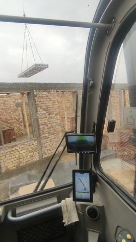 The view from inside a vehicle on a cloudy day captures construction activities outside. Through the windshield, a partially-constructed brick building is visible. A large object, possibly a slab or structure, is suspended by chains from above, against a gray sky. Inside the vehicle, two small screens are mounted on the dashboard. One displays what appears to be a live feed, while the other shows a map or tracking application. Wipers are positioned across the windshield, and a metal support bar is visible on the left side. The overall scene suggests construction work in progress, as viewed from the operator's cab of a crane or similar equipment.