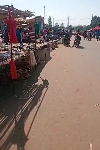 The media file captures a bustling outdoor market scene in Ngenyilel ward, Kenya. The time of day appears to be midday, indicated by the bright sunlight casting distinct shadows. The weather is sunny and clear.

The market is set up along a dirt road, with stalls lining one side. These stalls are made of simple wooden structures covered with tarps and corrugated metal, displaying a variety of goods. Visible items include clothing (shirts, dresses), footwear (sandals, shoes), and what appear to be household items or crafts. Bags of produce or other goods are also present, piled on the ground.

Several people are actively engaged in the market activities. Vendors are seen attending to their stalls, and shoppers are browsing the merchandise. Some individuals are standing, while others are walking or gathered in small groups. Two motorcycles are on the road, with one rider and passenger on the move, suggesting transportation is a key element of the market's accessibility. The general atmosphere appears lively and busy, characteristic of a typical marketplace.

There are no discernible texts visible in the media file. The location is identified as Ngenyilel ward, Kenya, which provides a geographical context for the scene. The overall impression is one of a vibrant community market contributing to local commerce and social interaction.