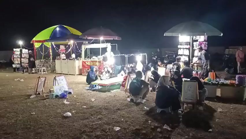This is a nighttime outdoor scene of a small market. Multiple stalls are set up, some with colorful umbrellas and lights. One stall appears to be selling small items, possibly toys or accessories, displayed on shelves. Another stall has a banner that says "MAKTRAK". Several people are gathered around the stalls, some sitting on low stools. A few people are standing and browsing the merchandise. The ground is covered in dry grass and scattered litter. The overall atmosphere seems to be that of a casual gathering or local market event.