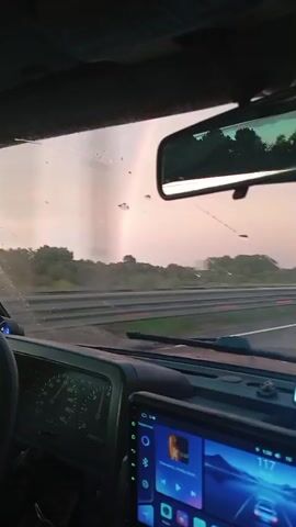 The video is taken from inside a vehicle, looking forward through the windshield. The dashboard and a touchscreen display are visible in the foreground. Outside, a road with multiple lanes and a divided barrier can be seen. Trees and foliage line the side of the road. In the sky, a faint rainbow is visible. The sky appears overcast with a pinkish hue, suggesting it might be dawn or dusk. The vehicle appears to be in motion.