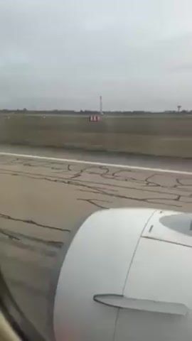 The video shows a view from an airplane window during what appears to be a takeoff roll on a runway. The wing and engine of the plane are visible in the foreground. The runway extends ahead, marked with lines. Beyond the runway, there is a grassy field with a tower and red and white striped poles in the distance. The sky is overcast and gray. The camera is relatively stable, with some minor vibration.