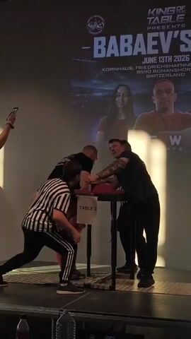 A referee in a black and white striped shirt and black pants stands between two men arm wrestling at a table. One of the men is wearing a black t-shirt and black pants, while the other is wearing a red garment. A woman in a pink shirt stands to the left, holding up a phone to record the event. Another person, wearing a black t-shirt and black pants, stands behind the referee. In the background, a large screen displays text that includes "KING OF THE TABLE PRESENTS - BABAEV'S" and "JUNE 13TH 2023 KORNHAUS, FRIEDRICHSHAFEN 88590 ROMANSHORN SWITZERLAND." The arm wrestling match is taking place on a raised platform with a black railing. There are two water bottles on the floor in the foreground. The lighting is dim, with a bright spotlight on the left side of the scene.