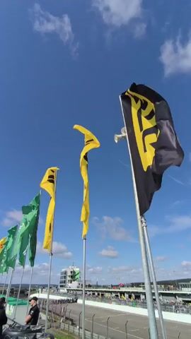 The video shows a series of flags waving in the wind against a backdrop of a clear blue sky with scattered clouds. The flags are predominantly yellow and black, with a few green ones at the left edge of the frame. A man in a black shirt is partially visible at the lower-left corner, likely a security or staff member. The flags are positioned along the edge of a racing track, with a low wall dividing the foreground from the track area. The track is visible in the distance, along with a control tower and some spectators. The flags appear to be promotional, featuring the logo of a racing organization. The wind is blowing strongly enough to make the flags billow and ripple.
