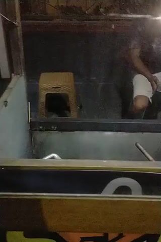A person is seen in a low-light setting, lying down. They are wearing a white shirt and shorts. There is a yellow object with a black interior next to them. In the background, there is a metal structure and some graffiti on a wall. The video appears to be recorded at night.
