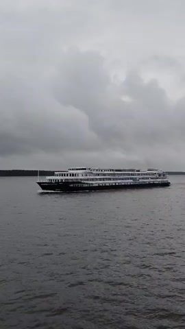 A wide shot shows a multi-decked passenger ship sailing across a wide body of dark water under a cloudy sky. The ship is dark with white decks and railings, and appears to be moving from left to right across the frame. The water is calm with small ripples. The sky is overcast with thick, gray clouds. The far shore is visible as a dark, tree-lined horizon.