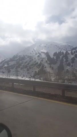 The video shows a snow-covered mountain landscape seen from a moving vehicle. The road is asphalt, with a metal guardrail separating it from the snowy terrain. Power lines are visible along the base of the mountain. The sky is overcast, with the sun occasionally breaking through the clouds. Trees dot the mountain slopes, partially covered in snow.