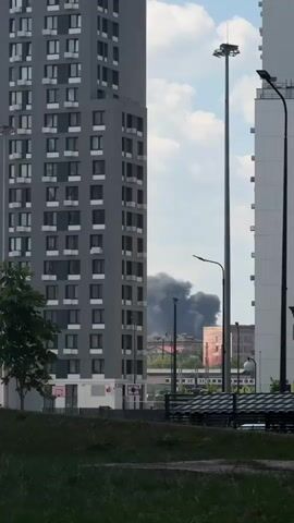 The video shows a view between two tall buildings. In the distance, a large plume of dark smoke rises into the sky. The smoke appears to be coming from an urban or industrial area. The sky is cloudy.