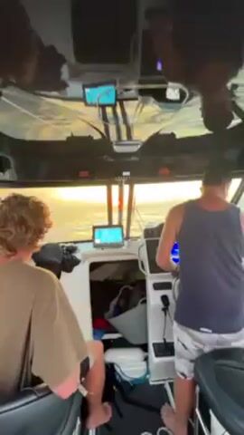 The video shows the interior of a boat moving on the ocean, with the sun setting on the horizon. There are two men in the boat, one with curly hair sitting in the passenger seat and another man steering. The camera angle moves to show the wake of the boat as it moves through the water. The sky is cloudy with patches of orange from the setting sun. The boat has various electronic navigation systems displayed on the dashboard.