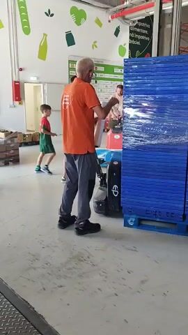 An older man wearing an orange shirt and grey pants is operating a blue pallet jack in what appears to be a warehouse or distribution center. He is moving a large stack of blue pallets wrapped in plastic. Another man, younger and wearing a white t-shirt, is standing near the stack of pallets, looking at the older man. In the background, there are shelves with boxes and signage on the wall depicting food items and graphics. The floor is concrete, and there is a metal ramp or edge in the foreground. The overall environment suggests a commercial setting for handling goods.