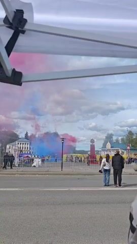 The video shows a public square where a vibrant display of red and blue smoke fills the air, creating a colorful backdrop against a partly cloudy sky. A barrier separates the viewer's location from the square, where people are gathered. Buildings and decorative structures are visible in the square. In the foreground, a couple stands near the edge of the road, observing the scene. Other people are scattered around the square, some walking and others standing. The overall atmosphere suggests a celebration or public event.
