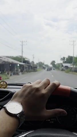 The video shows a first-person perspective of a person driving a car. The driver's hands are visible on the steering wheel. The road ahead is visible through the windshield, with other cars in the distance. The scenery outside the car consists of houses, trees, and utility poles. The sky is cloudy. A watch is visible on the driver's left wrist. A small ornament is attached to the dashboard.