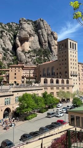 The video presents an eye-level, daytime shot of the Montserrat Monastery complex in Spain, situated against a backdrop of jagged, mountainous terrain under a clear, blue sky. The monastery is composed of various stone buildings, including a prominent square tower, built with a warm-toned material. The architecture features archways and numerous windows. Trees dot the courtyard, and people are visible strolling along the walkways. In the foreground, a street scene features parked cars and vans, a café (labeled with the sign "Caffè"), and pedestrians. The scene is well-lit, accentuating the natural colors of the buildings and landscape. The mountain serves as a dramatic backdrop, its unique rock formations dominating the skyline.