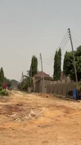 The video shows a shot of a residential street in what appears to be a developing area. The road is unpaved and dusty, with a few homes visible behind walls and fences. The houses appear to be built from concrete blocks and have roofs made of tile or corrugated metal. Power lines run along the street, supported by wooden poles. There is a small drainage ditch running alongside the road. The sky is a hazy blue.