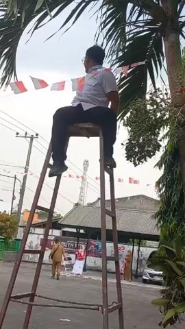 A man sits on a tall metal ladder with a seat attached. He is wearing a light-colored polo shirt, dark pants, and dark shoes. He is looking up and to his right. Red and white pennant flags are strung across the background. In the background, there are buildings and people, including a person in a tan uniform and a person in red and white attire. The scene appears to be outdoors with some trees visible.