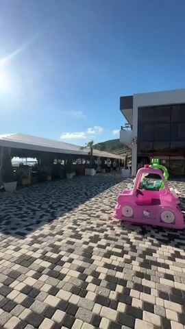 A bright, sunny day unfolds in this video, filmed outdoors. The foreground features a patterned walkway made of interlocking rectangular stones in shades of grey and white. To the right, a pink toy car with a green roof and a steering wheel is parked on the walkway. Beyond the walkway, a white-tented structure, possibly an outdoor dining area or cafe, is set up. Tables and chairs are visible beneath the tent, and several potted plants add greenery. People are seen walking and standing under the tent and in the distance. A building with large windows is visible on the right side of the frame, behind the toy car. In the background, the ocean can be seen, with a clear blue sky overhead. The sun is high in the sky, casting bright light and lens flare.