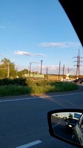 The video shows a view from inside a moving vehicle, looking out the passenger side window and into the side mirror. The landscape outside consists of a road, a grassy embankment, trees, and a clear blue sky with scattered clouds. In the distance, there is a train track with a train on it and several large power line towers. The side mirror reflects the road behind the vehicle, showing other cars on the road. The lighting suggests it is late afternoon or early evening.
