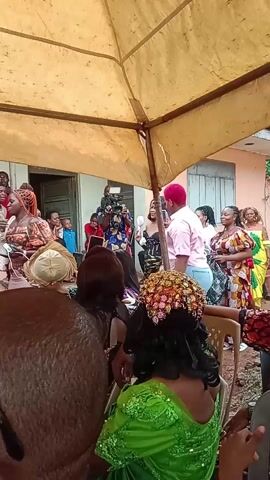 A group of people are gathered under a tan awning, appearing to be at an event. Most of the people visible are women dressed in various styles, many in colorful African prints. Some are standing and dancing near speakers, while others are seated. A man with a camera is visible, suggesting the event is being recorded. There's a building in the background, and the setting appears to be outdoors. Several men are seated in the foreground, observing the activities.
