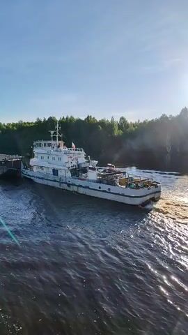 The image displays a wide-angle view of a white tugboat actively pushing a long, dark-colored barge along a river. The scene is bathed in bright sunlight under a clear, blue sky, suggesting either morning or late afternoon. The water exhibits a deep blue-green hue, with a prominent, sun-dappled wake trailing behind the moving vessels, indicating their forward momentum.

The tugboat is the central subject, a robust white vessel with a multi-tiered superstructure. Details such as railings and various pieces of equipment are visible on its deck. A small flag, possibly the Russian flag, is flown from a mast on the vessel's upper level. On the starboard side of the tugboat's hull, the number "738" is clearly legible, serving as a vessel identification mark. The tugboat is positioned at the stern of the barge, propelling it through the water.

The river is wide and stretches into the distance, flanked by dense, green forests that line both banks. The sunlight creates sparkling reflections on the water's surface, particularly in the disturbed wake. The overall setting is serene and natural, likely a waterway near Annensky Most, Russia. The bright, clear weather contributes to a peaceful atmosphere. No individuals are explicitly visible in the frame.