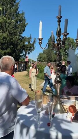 The video shows an outdoor wedding reception with guests mingling. A bride in a white dress and a groom are visible, along with other attendees dressed in casual attire. A golden candelabra stands prominently in the foreground, with champagne glasses and flower petals scattered on a nearby table, suggesting a celebratory atmosphere. The scene is set in a garden with trees and bushes in the background, under a clear blue sky. The camera appears to be handheld, capturing a candid moment of the event.