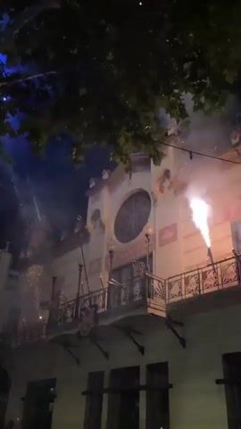 This video shows a building at night with fireworks being set off in front of it. The building has a balcony and is decorated with ornate details. There are trees in the foreground, obscuring some of the view. The fireworks are creating bright trails of light and sparks against the dark sky. Some smoke is also visible, indicating the combustion of the fireworks. It is difficult to see any people clearly due to the low light and distance. The overall atmosphere suggests a celebration or event taking place.