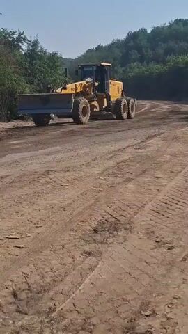 A yellow grader with the brand name SDLG visible on its side is operating on a dirt road. The grader has a large blade at the front and multiple wheels. The road appears to be under construction or maintenance, with loose dirt and tire tracks visible. Lush green trees and vegetation line the side of the road. The sky is clear and blue, suggesting a sunny day. In the background, there are hints of more trees and possibly a slight incline. Two figures, likely workers, are visible near the front left of the grader. One is wearing a light-colored shirt and dark pants, and the other is wearing a darker shirt and lighter pants. The grader is in motion, and its blade is actively engaged in leveling the dirt. The camera is positioned low to the ground, capturing the scene from a perspective slightly behind and to the side of the grader.