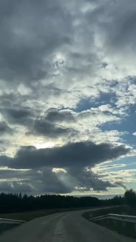 The video shows a view from inside a vehicle traveling on a gravel road. The road curves to the right in the foreground. On the left side of the road, there is a grassy field leading to a dense line of trees. The sky is filled with dramatic, grey and white clouds, with patches of blue sky visible. Sunlight is breaking through the clouds in some areas, creating beams of light. The overall impression is of a cloudy but bright day, possibly late afternoon. The camera is stationary relative to the interior of the vehicle, and the view is mostly of the exterior scenery.