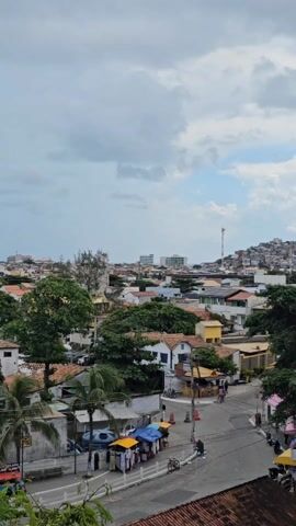 The camera pans across an overview of a town with white buildings, trees, mountains in the background, a street with vendor stalls, and a beach with many boats. The weather appears to be mostly cloudy.