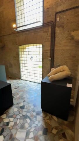 The video shows the interior of a room with ancient Roman artifacts. The walls are made of exposed brick and textured plaster. The floor is covered in a mosaic of small, colorful tiles. There are several sculptures on display, mostly torsos and heads, placed on black pedestals. A doorway leads to another room with more sculptures and a man standing near the entrance. There are windows with bars on them, allowing natural light to enter the space. A sign with text is visible on one of the walls.