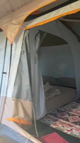 The video appears to be a personal vlog or travel diary documenting a camping trip in a lush, green environment.

**Subjects:**
*   **Objects:** The primary objects are a large tent with multiple windows and doors, camping furniture (chairs, possibly a mattress or sleeping pad), a lantern, and a woven rug. The tent interior is visible, showing a partitioned sleeping area with a bedroll and a rug. The exterior scene includes a wooden walkway or deck, a railing, and a suspension bridge in the background.
*   **Scene/Setting:** The setting is a campsite nestled within a dense forest or jungle. Tall trees with thick foliage dominate the background, suggesting a natural, somewhat wild environment. The presence of a wooden walkway and a suspension bridge hints at a more developed campsite or a nature reserve.
*   **People:** One person is partially visible in the foreground, seated on a camping chair, with their back to the camera. Their hair and clothing suggest a relaxed, casual style.

**Activity or Event:**
The media captures moments of a camping trip. It transitions from an interior view of the tent, showcasing its amenities and setup, to exterior shots of the surrounding natural landscape. The activity appears to be leisurely, focused on enjoying the camping experience and the natural surroundings.

**Notable Details:**
*   **Time of Day:** The lighting suggests it could be late afternoon or early evening, with the sky appearing somewhat overcast or dusky. The lantern being lit in one shot reinforces this idea.
*   **Weather:** The dense foliage and the subdued lighting might indicate a humid or slightly overcast day, common in tropical or subtropical regions.
*   **Location Cues:** The lush vegetation, specific types of trees (possibly pines or similar), and the style of the wooden structures (walkway, bridge) are strong indicators of a tropical or sub-tropical natural environment. The mention of "Batu Alang, Indonesia" provides a definitive geographical context.
*   **Emotions:** The overall mood conveyed is one of relaxation, peace, and connection with nature. The interior shots emphasize comfort and preparedness for the outdoors, while the exterior shots highlight the beauty and tranquility of the natural setting.

**Visible Text:**
No legible text is visible in the media.