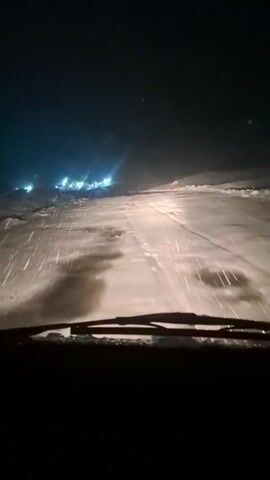 The video shows a car's view from the inside, driving at night on a snowy road. Snowflakes are heavily falling, lit up by the car's headlights and a distant cluster of lights in the background. The road is covered in snow, and the windshield wipers are actively clearing the snow. The rest of the car's interior is dark, only the lower part of the windshield and the wipers are visible.