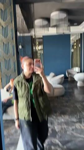 A woman stands in front of a full-length mirror taking a selfie with her phone. She has reddish-brown hair pulled back in a bun, wears a green puffy vest over a black t-shirt, and dark baggy pants. She holds a phone with a patterned case in her right hand, facing the screen, and makes a kissy face. The environment includes decorative gold and blue wall panels behind her, modern white chairs, hanging circular lights, and dark, patterned floor tiles. Another person is barely visible in the background, seated on a sofa.