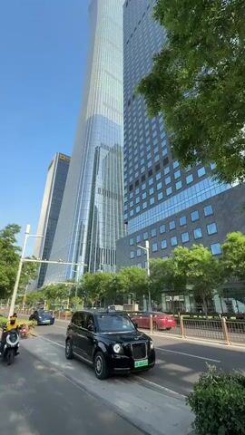 The video depicts a modern urban landscape, likely in a major city with high-rise buildings. The sky is clear and blue. A black London taxi drives along a wide street with trees lining the sidewalk. Another car, a blue sedan, follows the taxi. A person on a small motorcycle or scooter is briefly visible at the start of the video, dressed in a yellow uniform. The architecture of the buildings is characterized by glass and steel, indicative of contemporary design. Overall, the scene conveys an impression of an advanced and prosperous urban environment.