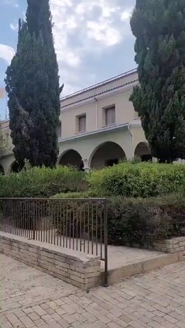 The video shows a pathway leading to a building with arched entrances, surrounded by lush greenery and tall cypress trees. A flag with a yellow emblem on a white background is visible on a flagpole to the left of the building. The sky is blue with scattered clouds. The pathway is paved with light-colored bricks.
