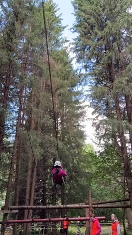 A person wearing a helmet and a pink jacket is ziplining through a forest. The person is suspended from a cable by a harness. Tall trees surround the zipliner. There are also some wooden structures visible below.