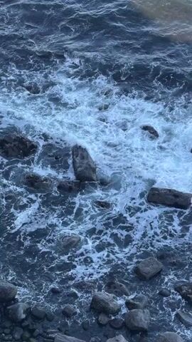 The video begins with a close-up shot of waves crashing over rocks in the ocean. The camera then pans up to reveal a rocky coastline, with a cliff dropping down to the ocean below. The water is a mix of deep blue and turquoise, and the sky is a clear, bright blue. There is a waterfall cascading down the cliff face into the sea. The coastline is rugged, covered with grass and rocks. The sun is shining, and the overall scene is one of natural beauty.