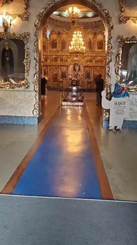The video shows the interior of an ornate church. A blue carpeted aisle extends from the foreground towards a golden iconostasis. Several icons are visible on the iconostasis, along with a large chandelier. Two people are standing near the iconostasis, one in profile facing the altar, the other facing away from the camera. A third person is partially visible on the left. In the foreground, a plaque with Russian text is mounted on a stand. The walls are decorated with framed images and ornate golden moldings. The lighting is dim, with the chandelier and several candles illuminating the space.