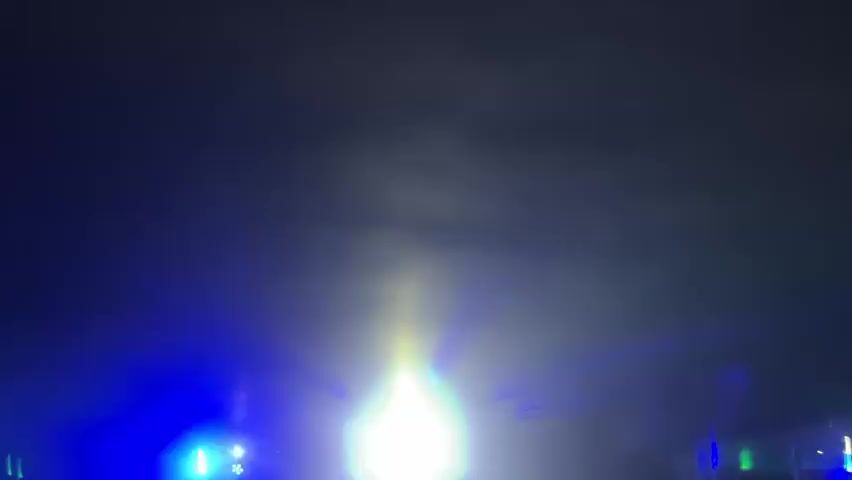 The video shows a dimly lit room with a ceiling that appears to be textured. There are bright spotlights of yellow and blue light shining down, creating beams that illuminate the smoke or haze in the air. A crowd of people, mostly silhouetted, are gathered in the room, facing the direction of the lights. Some individuals have their hands raised. The atmosphere suggests a party or a concert. The camera is positioned behind the crowd, looking towards the front of the room where the lights are originating. The lighting changes from blue and yellow to green and then back to bright white spotlights.