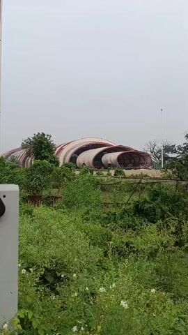 A wide shot shows a large, futuristic building with a distinctive curved roof structure, resembling a series of interconnected shells or tubes. The building is situated on a slight rise and surrounded by lush green vegetation, including trees and bushes. In the foreground, there is dense foliage. The sky is overcast. The overall impression is of an abandoned or under-construction modern architectural site.