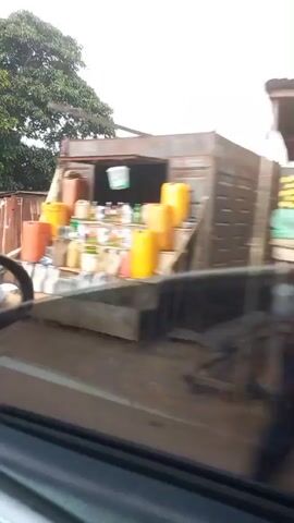 A low-angle, moving video captures a street scene with various buildings and vehicles. The video begins with a blurry image of a white plastic bag on the ground in front of corrugated metal structures. Then, a blue car is seen passing by, with a person in an orange outfit standing next to it. A silver car follows closely behind. The video pans to reveal a two-story building with a sign that reads "OBAA BUSINESS ENTERPRISE". In the foreground, a car mirror reflects the passing scene. Further down the road, a house with a light-colored roof is visible, along with stacks of white bags. The sky is overcast.