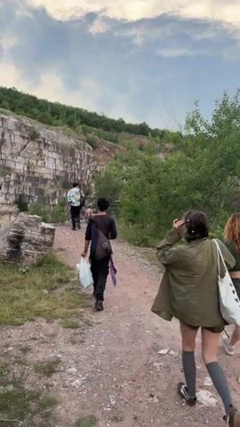 Five people walk in single file along a dirt path next to a large stone quarry wall on the left. The person at the front-left is a female wearing an oversized olive green shirt, dark shorts, and long grey socks, carrying a white tote bag. To her right, another female wears a green crop top and shorts, carrying a green object. Behind them, a male in a black shirt and black pants carries multiple white plastic bags. Further back, another male in a white patterned shirt and black pants carries a backpack. Green trees and shrubs line the path to the right, and a partially cloudy sky is visible overhead.